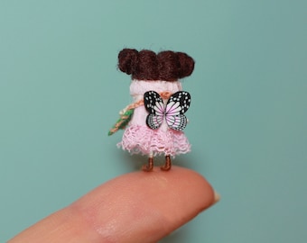 Puede incluir: Una pequeña muñeca rosa con cabello castaño y un ala de mariposa. La muñeca lleva un vestido rosa con encaje y está de pie sobre un dedo.