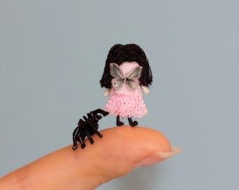 Puede incluir: Una pequeña muñeca con cabello negro y un vestido rosa está de pie sobre un dedo. La muñeca tiene una mariposa en la espalda. Una araña de plástico negra está arrastrándose por el dedo.
