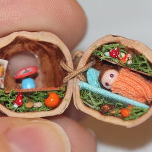 Op de afbeelding: Een gedetailleerd miniatuur diorama in een walnootschil. De ene helft toont een slapende pop onder een oranje deken, de andere een scène met een weegschaal, paddenstoelen en een vogelafbeelding. De twee helften zijn verbonden door een bruin touwtje.