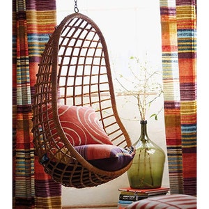 May include: A brown wicker hanging chair with a red and white patterned cushion. The chair is hanging in front of a window with striped curtains.