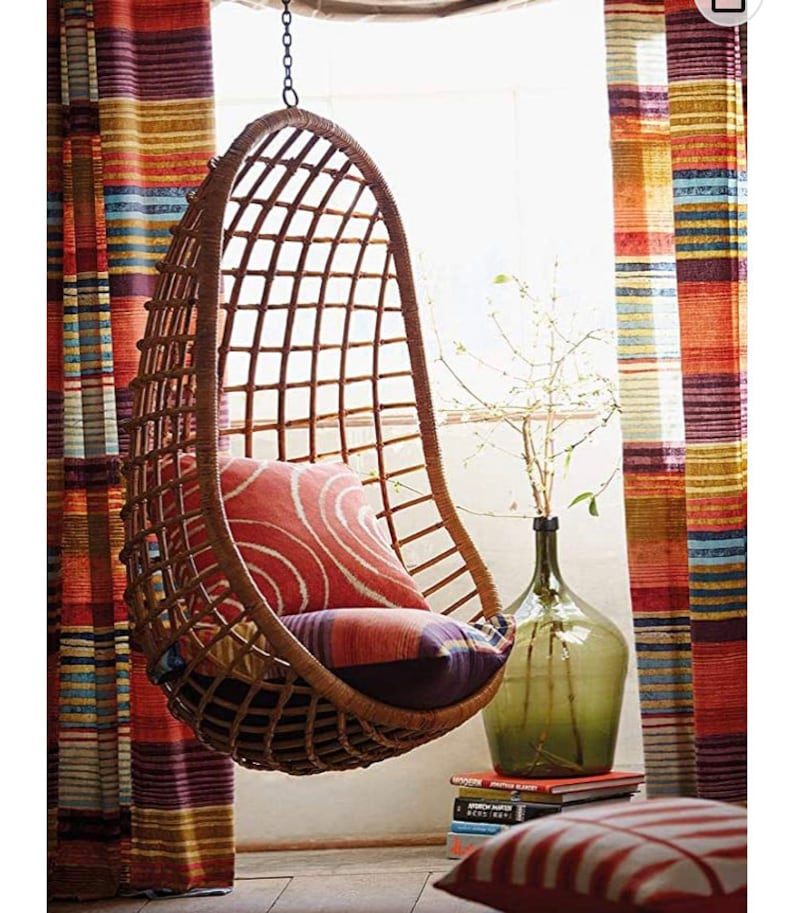 May include: A brown wicker hanging chair with a red and white patterned cushion. The chair is hanging in front of a window with striped curtains.