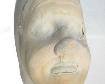 May include: A pale, textured mask of a human face with closed eyes, a prominent nose, and an open mouth. The mask has visible wrinkles and is likely made of a flexible material. The text "FX Warehouse" is visible at the bottom.