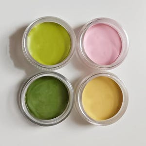 May include: Four small, clear jars filled with colorful cosmetic products. The jars contain shades of green, pink, and yellow. The products appear to be creamy or waxy in texture, and are arranged in a square formation.