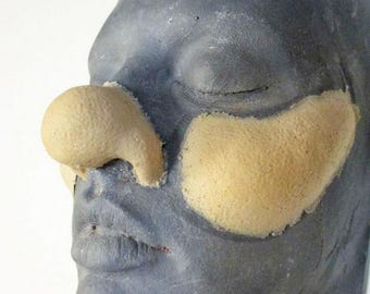 May include: A grey face with closed eyes, featuring prosthetic nose and cheek pieces. The nose and cheek pieces are a light beige colour, contrasting with the grey skin tone. The prosthetic pieces have a textured surface.