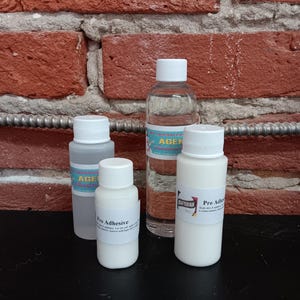 May include: Four bottles of craft supplies are displayed against a brick wall. The bottles vary in size and contain clear and white liquids. One bottle has a label that reads "Pro Adhesive". Another bottle has a label that reads "AGENT".