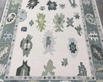 May include: A rectangular area rug with a cream-colored central field and a teal border. The rug features a repeating pattern of abstract floral and geometric shapes in shades of green, gray, and black. The rug's design is symmetrical.