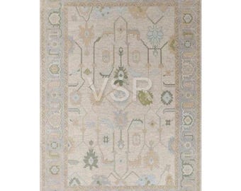 May include: Rectangular area rug with a beige central field and a detailed border. The rug features a symmetrical design with floral and geometric patterns in shades of green, blue, and tan. The rug has a distressed, vintage look.