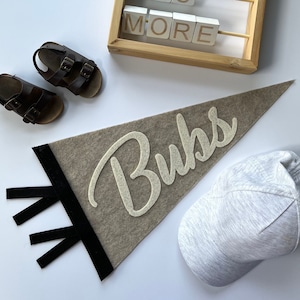 May include: A gray felt pennant with the word "Buks" in white felt letters. The pennant is hanging from a black ribbon. There is a gray baseball cap, a pair of brown sandals, and a wooden frame with the words "Love You More" in white letters on wooden blocks.