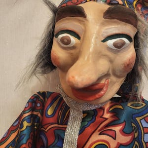 May include: A close-up of a handmade puppet with a painted face, gray hair, and a colorful patterned hat and tunic. The puppet has a long nose, red lips, and a silver scarf. The tunic is a mix of red, blue, yellow, and orange.