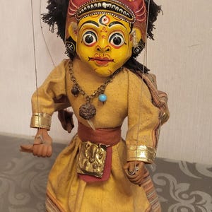 Puede incluir: Títere tradicional con rostro amarillo pintado, cabello negro y tocado rojo. El títere viste una túnica amarilla con ribetes dorados y un collar. Un artefacto cultural, probablemente para la narración o el entretenimiento.