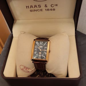 Könnte beinhalten: Eine rechteckige goldfarbene Armbanduhr mit schwarzem Zifferblatt, weißen arabischen Ziffern, einer Tages- und Datumsanzeige und einem braunen Lederarmband. Die Uhr liegt auf einem cremefarbenen Kissen in einer braunen Box mit HNC HAAS & CIE SINCE 1848.