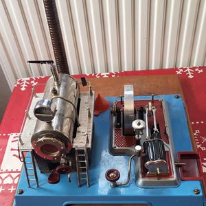 Könnte beinhalten: Ein antikes Modell einer Dampfmaschine mit silbernem Kessel, Manometer, hohem braunen Schornstein und komplizierten mechanischen Komponenten. Die Maschine ist auf einer blauen Basis mit roten Akzenten und einer Holzplattform montiert.