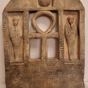 Può includere: Un rilievo in pietra rettangolare con una parte superiore ad arco. Il rilievo presenta una finestra centrale con un simbolo ankh, affiancato da due figure in pose mummificate. Le figure sono circondate da geroglifici. La parte superiore del rilievo presenta uno scarabeo e due figure sedute su sedie.