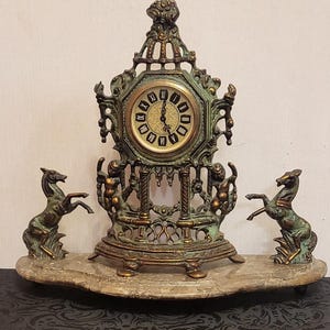 Op de afbeelding: Een schoorsteenklok met een groen en goudkleurig metalen frame met sierlijk krulwerk, rustend op een marmeren voet. Twee steigerende paarden flankeren de centrale wijzerplaat met zwarte Romeinse cijfers op een crèmekleurige achtergrond.