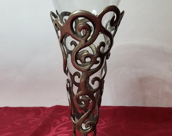 Könnte beinhalten: Eine silberne Metallvase mit einem klaren Glaseinsatz und einem dekorativen, wirbelnden Design. Die Vase hat eine breite Basis und eine schmale Öffnung.