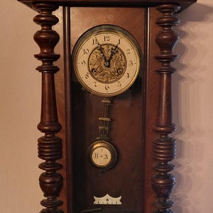 May include: A dark wood wall clock featuring a gold-colored lion figurine on top. The clock face has black Arabic numerals and a central gold-toned decorative plate. A pendulum with a circular brass disc marked with the letters R and A hangs below.