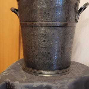 Puede incluir: Un cubo de vino de metal gris oscuro con un patrón floral detallado. El cubo tiene dos asas curvas y se asienta sobre una superficie estampada gris oscuro. Es un artículo decorativo.