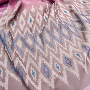 May include: A folded textile with a pink and mauve color scheme. The fabric features a repeating geometric pattern in white, blue, and pink. The design includes diamond and zigzag shapes, creating a textured appearance. The edges of the fabric are finished with a contrasting pink border.