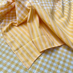 May include: Yellow and white gingham and striped fabric. The fabric is folded and draped, showing both the gingham and striped patterns. The colors are bright and the fabric appears to be cotton or a similar material.