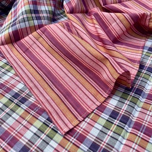 May include: Close-up of a folded fabric with a plaid and striped pattern. The plaid features a mix of red, purple, green, and white. The striped section has vertical lines in shades of red, pink, and orange. The fabric appears to be cotton.
