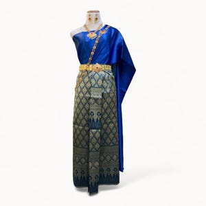 May include: A blue and gold traditional Thai dress with a patterned skirt and a matching shawl. The dress is decorated with gold jewelry, including a necklace, earrings, and a belt.