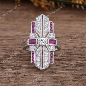 Peut inclure: Une bague en argent avec une grande pierre centrale ronde et transparente, entourée de pierres plus petites et transparentes. La bague est également ornée de pierres rectangulaires rouges serties le long de la bande.