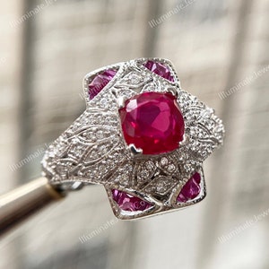 May include: A silver ring with a large, red, cushion-cut gemstone surrounded by smaller, white gemstones. The ring has a detailed, floral design.