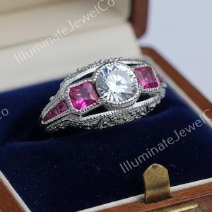 May include: A silver ring with a large, round, clear center stone and smaller, rectangular pink stones. The ring is set in a decorative band and is displayed in a wooden jewelry box with a blue velvet interior.