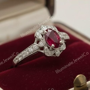 Könnte beinhalten: Ein silberfarbener Ring mit einem ovalen Rubin als Mittelstein, umgeben von einem Halo aus kleinen Diamanten. Das Band ist ebenfalls mit Diamanten besetzt. Der Ring wird in einer Vintage-Schmuckschatulle mit rotem Samtfutter präsentiert.