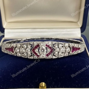 Può includere: Un bracciale in stile Art Déco con un diamante centrale e dettagli in rubini, presentato in un cofanetto di velluto blu. Il bracciale presenta un design geometrico con un motivo centrale a forma di occhio. La scatola è impressa con il nome "YADRUM".