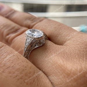 May include: A silver engagement ring with an oval-shaped center stone and intricate filigree detailing. The ring is set on a person's finger.