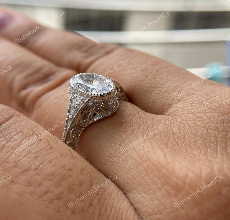 May include: A silver engagement ring with an oval-shaped center stone and intricate filigree detailing. The ring is set on a person's finger.