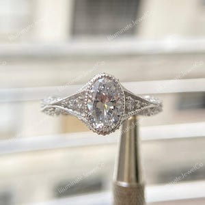 May include: A silver engagement ring with an oval-shaped diamond set in a delicate, ornate setting. The ring features a halo of smaller diamonds surrounding the center stone.