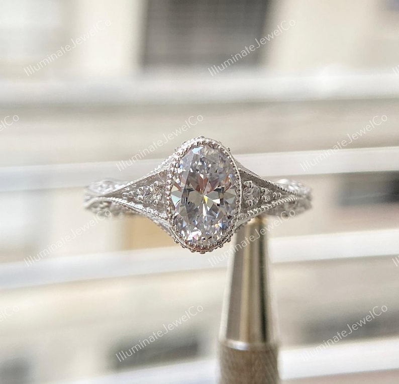 May include: A silver engagement ring with an oval-shaped diamond set in a delicate, ornate setting. The ring features a halo of smaller diamonds surrounding the center stone.