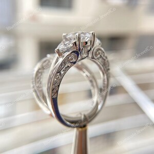 May include: A silver engagement ring set with a pear-shaped center stone and two smaller round stones on either side. The band is intricately engraved with floral designs.