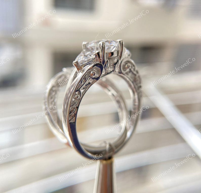 May include: A silver engagement ring set with a pear-shaped center stone and two smaller round stones on either side. The band is intricately engraved with floral designs.