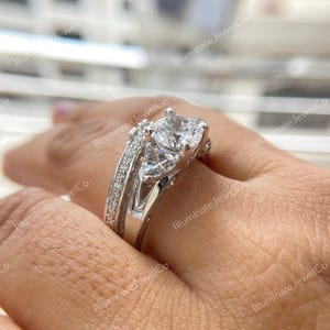 May include: A silver three-stone engagement ring with a large round center stone and two smaller pear-shaped stones on either side. The ring is set on a band with a delicate, intricate design.