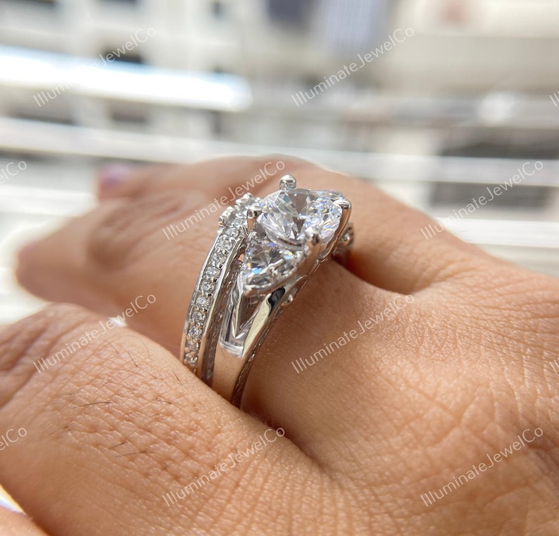 May include: A silver three-stone engagement ring with a large round center stone and two smaller pear-shaped stones on either side. The ring is set on a band with a delicate, intricate design.