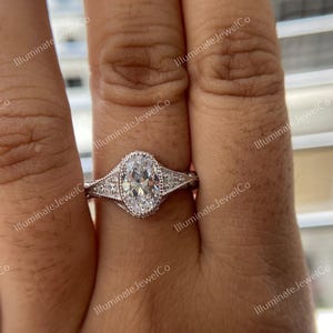May include: A silver engagement ring with an oval-shaped center stone and smaller stones set in the band. The ring is on a finger.