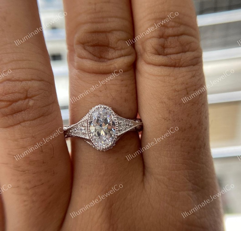 May include: A silver engagement ring with an oval-shaped center stone and smaller stones set in the band. The ring is on a finger.