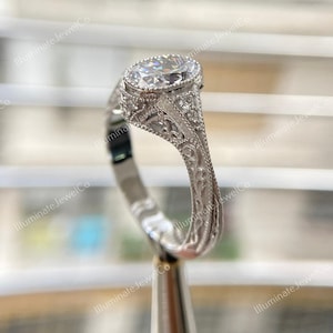 May include: A silver engagement ring with a large oval-shaped diamond set in a filigree design. The ring is on a display stand.