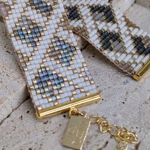 Op de afbeelding: Een witte en grijze kralen armband met een gouden sluiting en een klein gouden label met de tekst "ISABELLA".