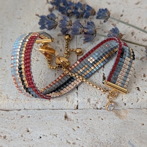 May include: A beaded bracelet with a gold clasp and chain. The bracelet has a pattern of red, black, gold, and grey beads. A small, clear charm hangs from the chain. Dried lavender sprigs are in the background.