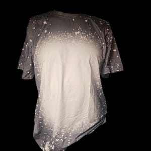 May include: A short-sleeved t-shirt with a unique design. The shirt transitions from a dark gray at the top to white in the center, with white splatter effects. The garment is displayed against a black background.
