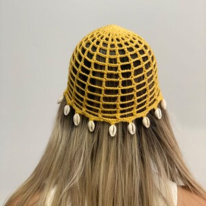 May include: A yellow crocheted head covering with a grid-like pattern. The head covering is adorned with small, white seashells along the bottom edge. The head covering is worn on a person with long, blonde hair.