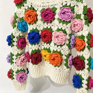 May include: A white crocheted sweater with colorful crocheted flowers. The sweater has a crew neck and long sleeves.