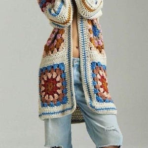 May include: A long, crocheted cardigan with a colorful geometric pattern. The cardigan is open in the front and has a relaxed fit. The model is wearing the cardigan over a pair of blue jeans.