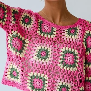Puede incluir: Un top de ganchillo rosa con un patrón floral en verde y amarillo. El top tiene mangas largas y un cuello redondo. El diseño de ganchillo es abierto, creando un efecto transparente. El top es de estilo crop.