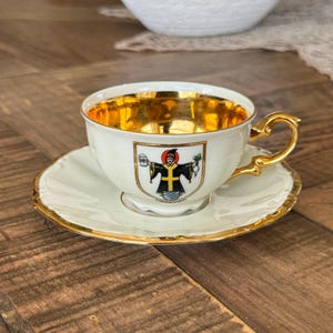 May include: A white teacup and saucer set with gold trim and a gold interior. The teacup features a crest with a figure in a black robe. The set is placed on a wooden surface.