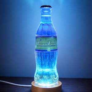 Op de afbeelding: Een blauw gloeiende flessenlamp met de tekst "Quantum". De lamp is gemaakt van een doorzichtige fles met een zwarte dop, verlicht door blauwe LED-lampjes. Hij staat op een ronde houten voet met een wit netsnoer.